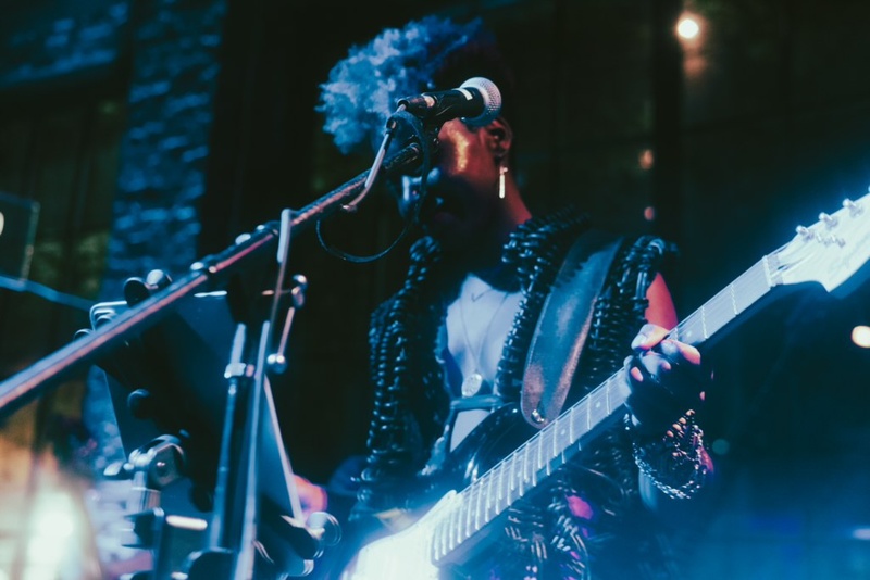  Shimmery stage lights on a Black woman with short hair holding her guitar and standing before a microphone 
