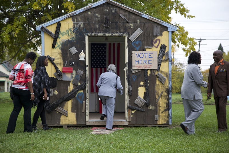 There is an outdoor shed with an opening that someone is walking into. There is an American flag inside and a sign on the outside that says VOTE HERE. 