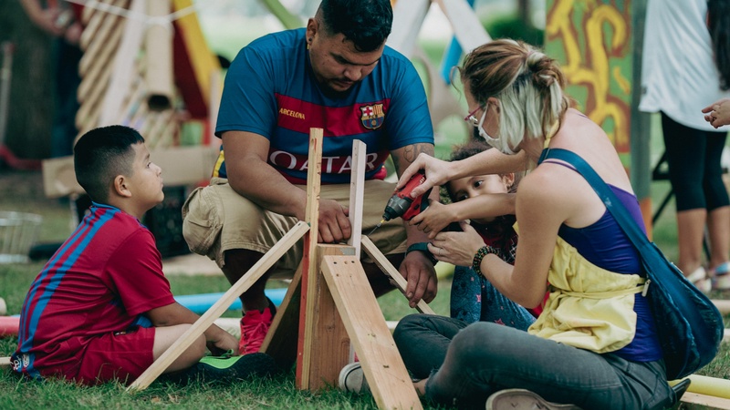  A group creates a small wood structure outdoors: A young girl presses the trigger of a power drill, Marya stabilizes the tool, the girl's father holds a board in place, and his son watches. Beyond them, various boards and objects are creatively installed. 
