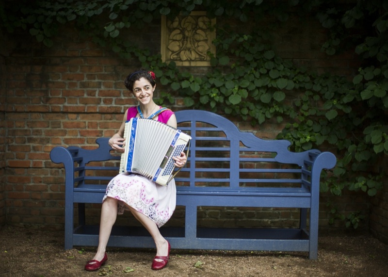  woman dressed in pink and red happily playing a blue accordion on a blue bench 