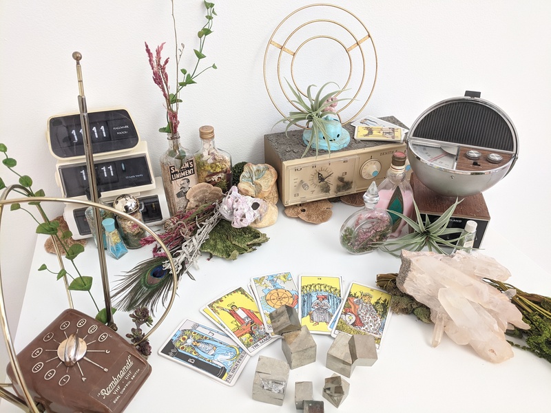  white pedestal with installation consisting of vintage radios and clock radios, vintage brown TV antenna, several vintage apothecary bottles filled with dried flowers, assorted crystals, tarot cards arranged in fanned half-circle 