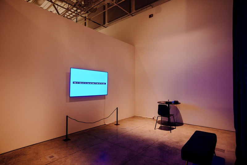  Photo of the gallery exhibition installation. It shows a large monitor on the wall across from a round table with audio gear on it. The table holds the customized controller, headphones, and headphone amplifier. The monitor has a bright blue background wi 