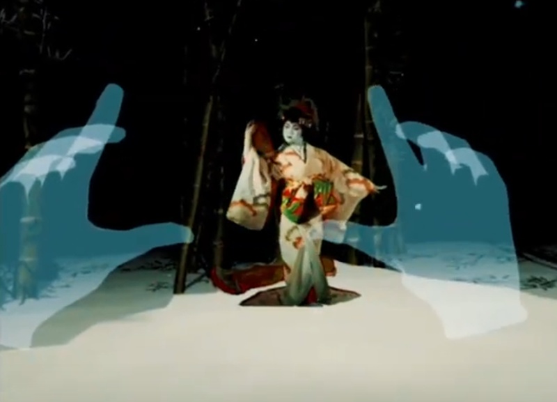  An Asian American woman dressed in traditional Japanese kinimo dancing in a dark room between two virtual hands. 