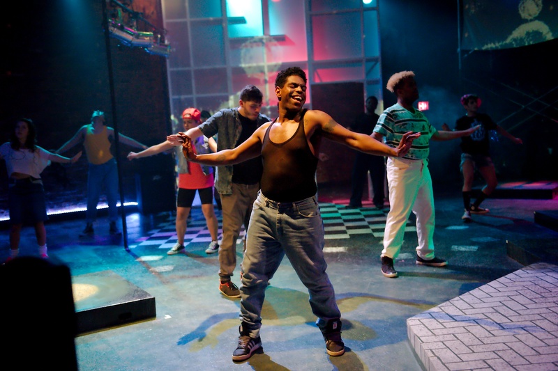  Man wearing tank top and jeans has a broad smile and arms outstretched in a dance move with others assembled behind him 