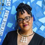 Black woman with long locs, black glasses, standing in front of a bright blue mural