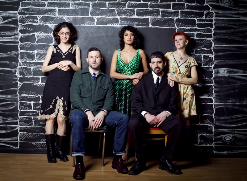  In this portrait, Ami stands in the center of her bandmates. She wears a green dress which pops on the black chalk wall background. In front of her are two people sitting and two people stand to the side of her. 