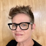 Picture of a white female 50 something with short spiky hair and chunky framed glasses, blue eyes, and a little grin.