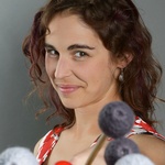 A white woman with wavy brown hair smiles slyly at the camera, holding a pair of percussion mallets out toward the viewer. She is wearing a red, black and white patterned blouse in front of a gray background.