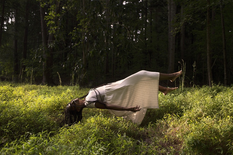  A Black woman with braids in a white cotton dress, floats on her back above a lush grass. 
