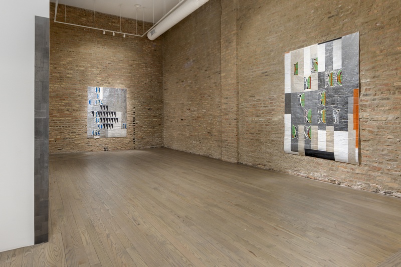  A wide-angle view of a gallery space with brick walls. Left of center, towards the back of the space and on the right wall another there are two large mixed media works that have a silvery reflective ground with colorful embroideries of fragmented letters 