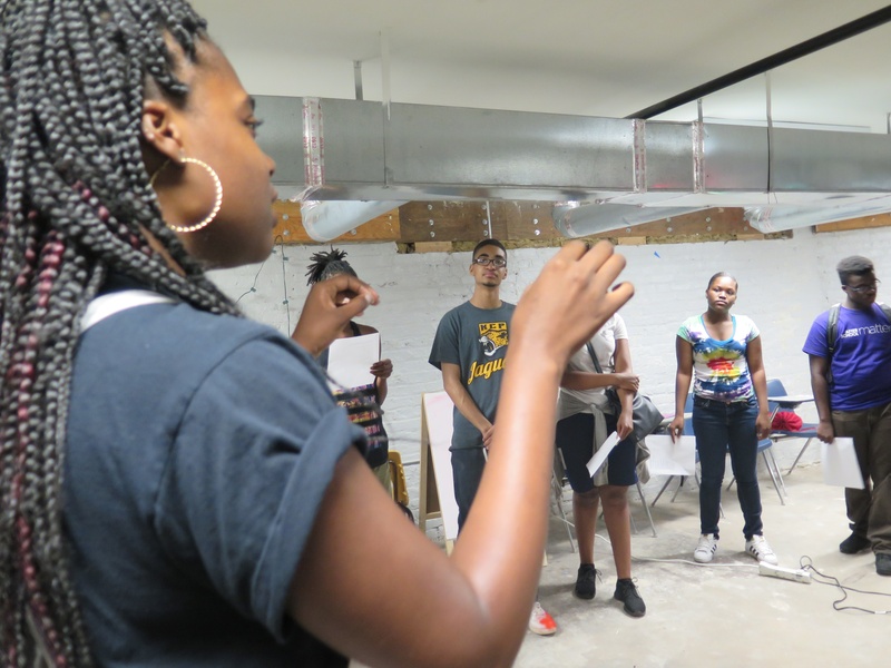  Black person with long braids at the left of the frame, addressing a group of youth 