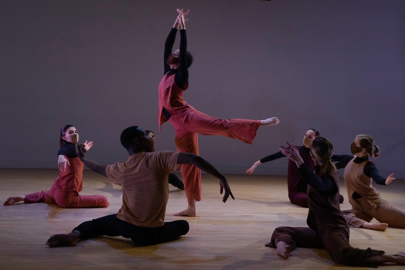  A group of dancers sit on a floor in a circle, surrounding another dancer standing on one leg with arms upstretched. 