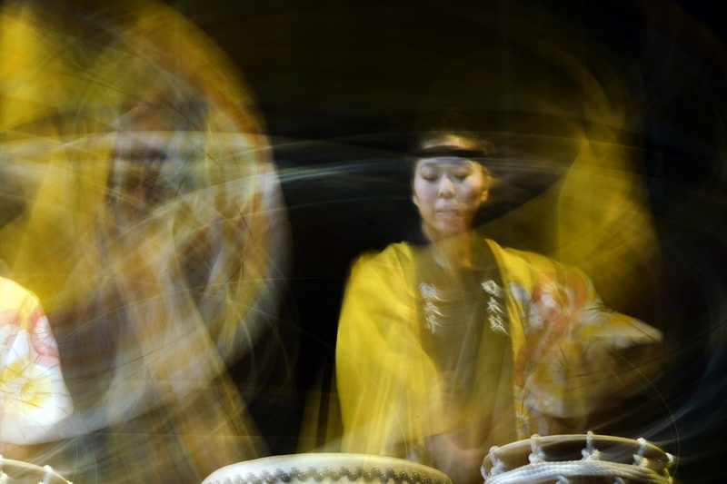  A master Taiko drummer, Kioto is dressed in yellow, gently swaying as she percusses two Taiko drums. She is sitting near a large Taiko drum that has dissolved in different colored textures and light. 