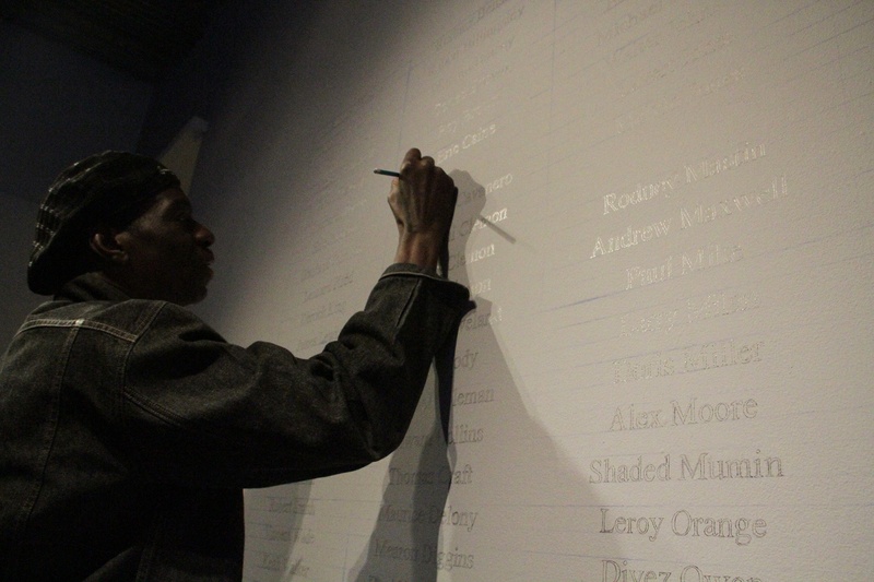 Torture survivor Darrell Cannon, a tall Black man in silhouette, writes his name on a large hand-drawn Wall of Names honoring survivors of police torture.