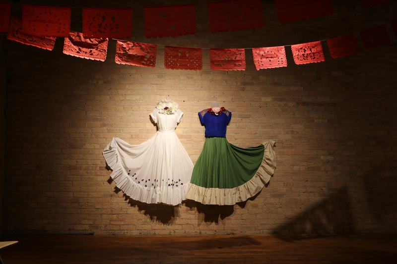  a brick wall with two dresses displayed. The left is white with small flowers on the skirt. The right is a blue top and green skirt with tan ruffle. They are replicas of the dresses in Frida Kahlo's "Dos Fridas" 