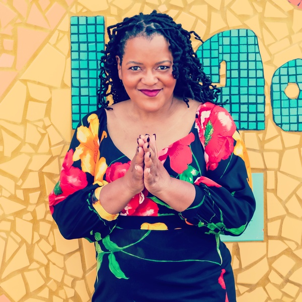  Black woman with hands together in flowered dress in front of yellow and green mosaic 