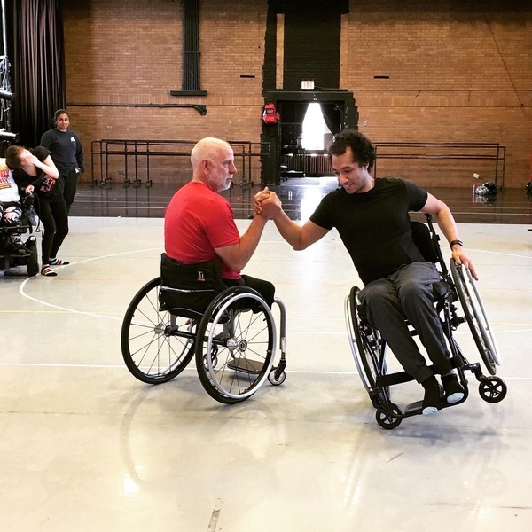  Two dancers in wheelchairs, side-by-side, gripping hands while one is tilted on one wheel 
