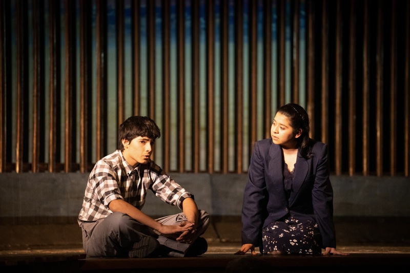  Two people sitting in a spotlight on the edge of a stage talking to each other. 