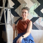 An image of Dianna Frid sitting on a chair in front of two works in progress that are only seen partially. A ladder is next to her. She is smiling softly and looking at the camera. She is also barefoot, but this is not visible because the image is cropped