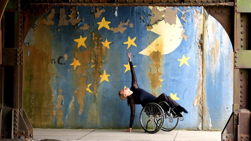  Ladonna tilted back in her wheelchair with one arm reaching towards a star on the nightime sky mural behind her 