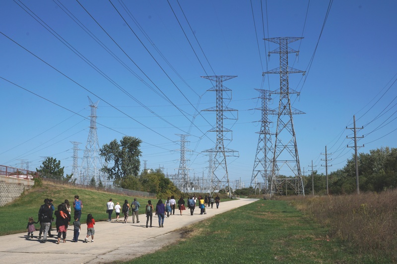  Faraway image of a larger group of people walking on a concrete path, approaching a cluster of giant metal electrical grid towers 