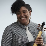 Photo of Ayriole, a brown skinned Black person wearing a black and white checked shirt, a white tie, glasses and dangly earrings. Their natural hair is styled in a sweep over their forehead with the rest pulled back. They are holding a viola and smiling.
