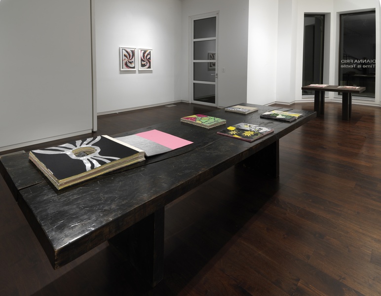  A dark brown table in the foreground of a gallery space. Five open one of a kind artist’s books of diverse colors and sizes are on the table. In the background there is a framed two-part mixed media work with embroidered text. The photo was taken at night 