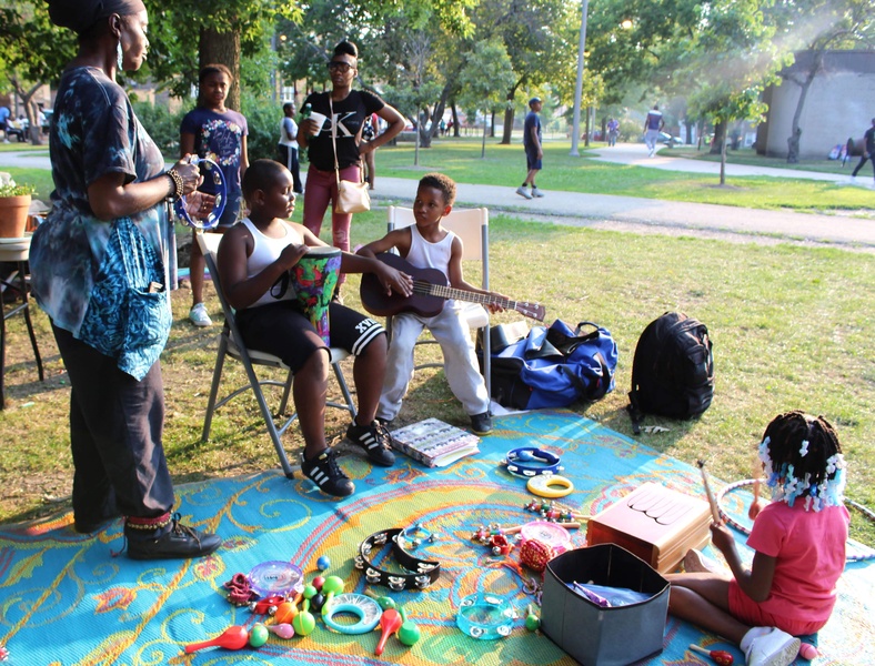  Black woman stands to the left instructing two young people how to play guitar instruments. 