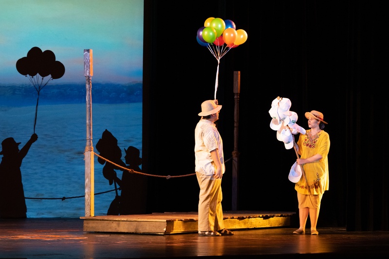  Two people standing under a spotlight on stage. One person is holding a bundle of colorful balloons and the other is holding a stick with hanging hats. 