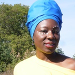 Black woman with smiling brown eyes, wearing a blue headwrap with face adorned with a gentle curving line of white dots around her eyes