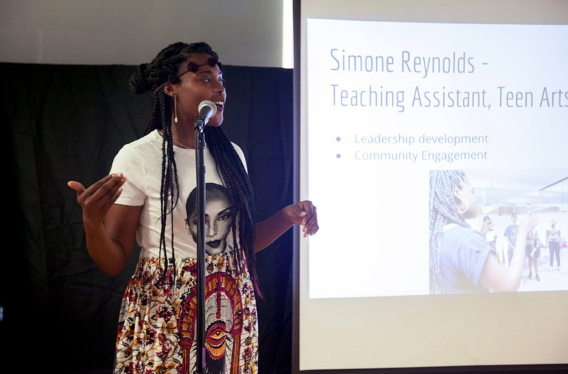  Black person with long braids stands speaking at a microphone in front of a screen that reads "Simone Reynolds, teaching assistant" 