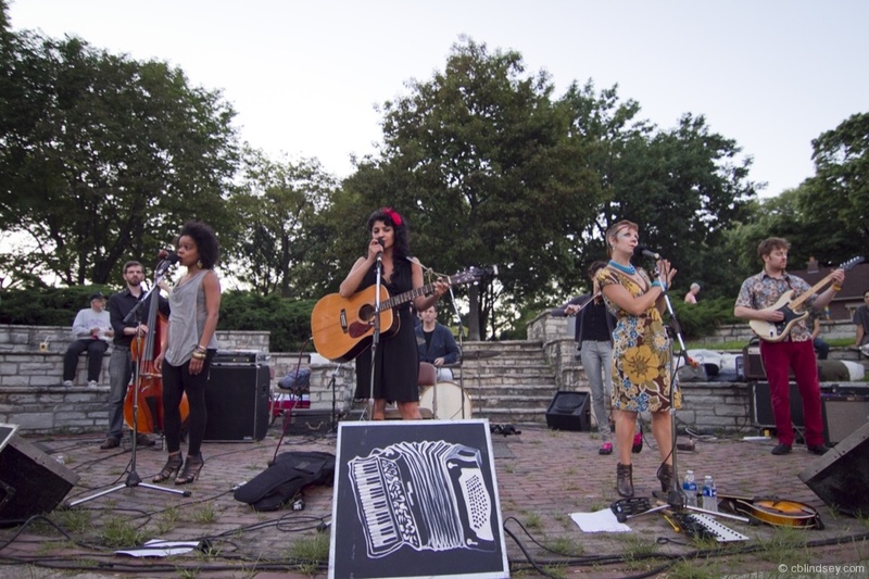  Ami performs outside surrounded by band members. They hold different instruments and have large trees behind them. 