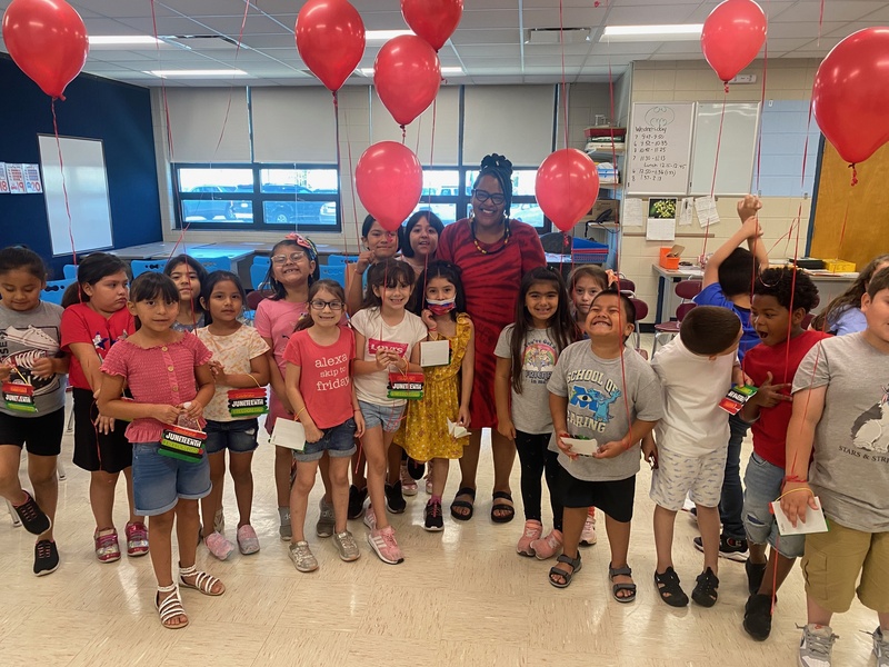  Classroom with glass windows, red balloons, several young children standing in a semi circle black woman in red dress in the center 