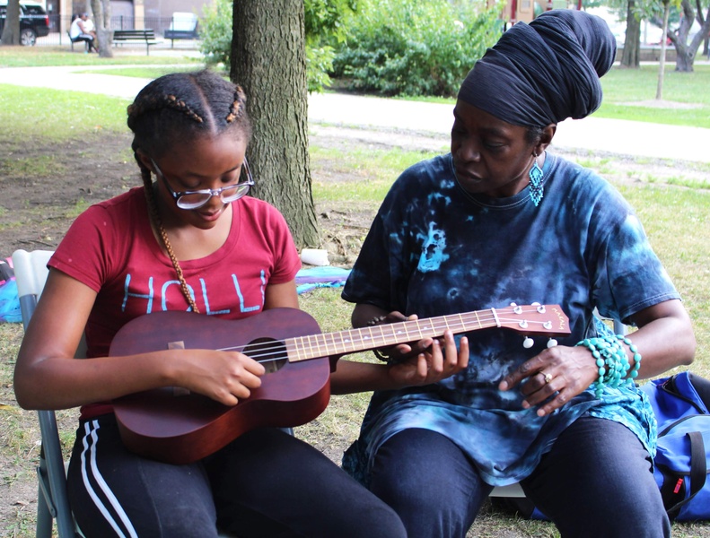  Two people sit beside each other. Person on the left holds brown ukulele while Black woman on the right instructs them how to play. 