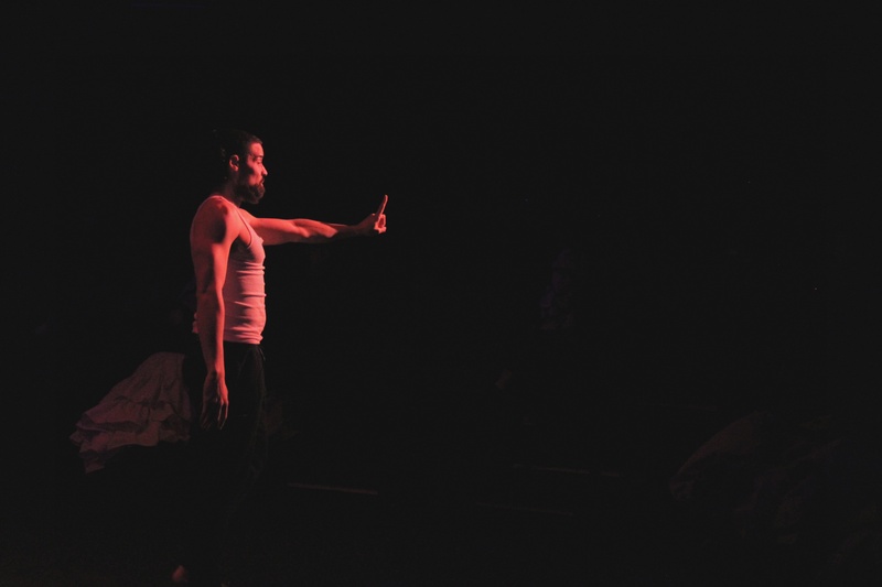  A light skinned femme in white under shirt and navy pants stands on a black stage under a red light, with their left index finger outheld. 