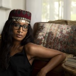 A Black woman with Glasses and a hat with jewels hanging over the forehead leans against a chair with her arm resting on it.