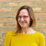 The photo shows Marya Spont-Lemus from the chest up and smiling slightly. Marya, a white person, is wearing pink glasses and a yellow sweater and has an asymmetrical haircut. She appears against a brick background. Photo by Gaby FeBland.