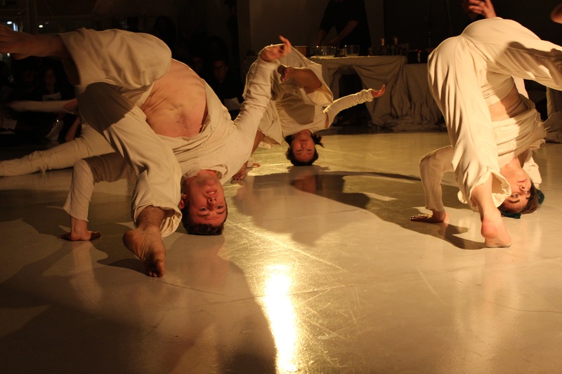  A White man, Asian woman, and White woman in loose cream colored costumes move through an inversion. The crowns of their heads support their axial weight while they move coronally in unison from their right limbs to left limbs. 