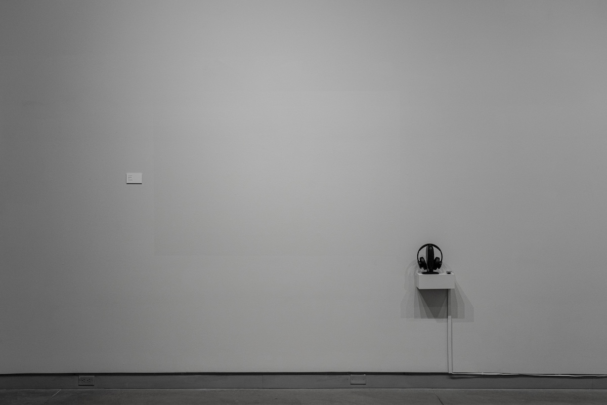 Image of a white gallery wall. On the right side of the wall is a small platform with headphones.