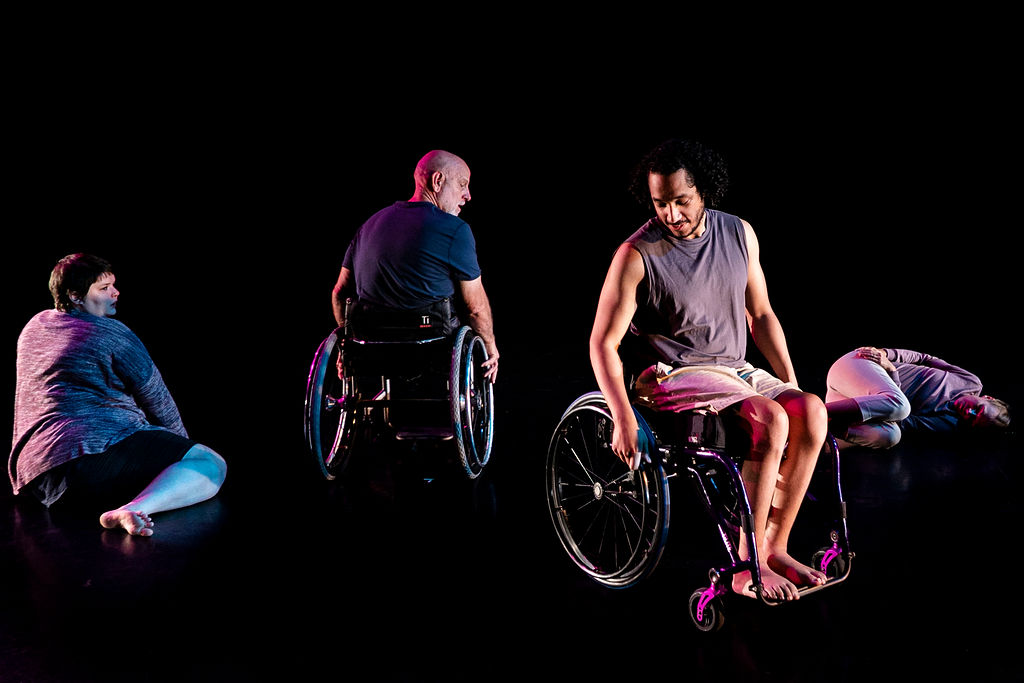 Four figures including two in wheelchairs are seated or paused in motion on a dance stage