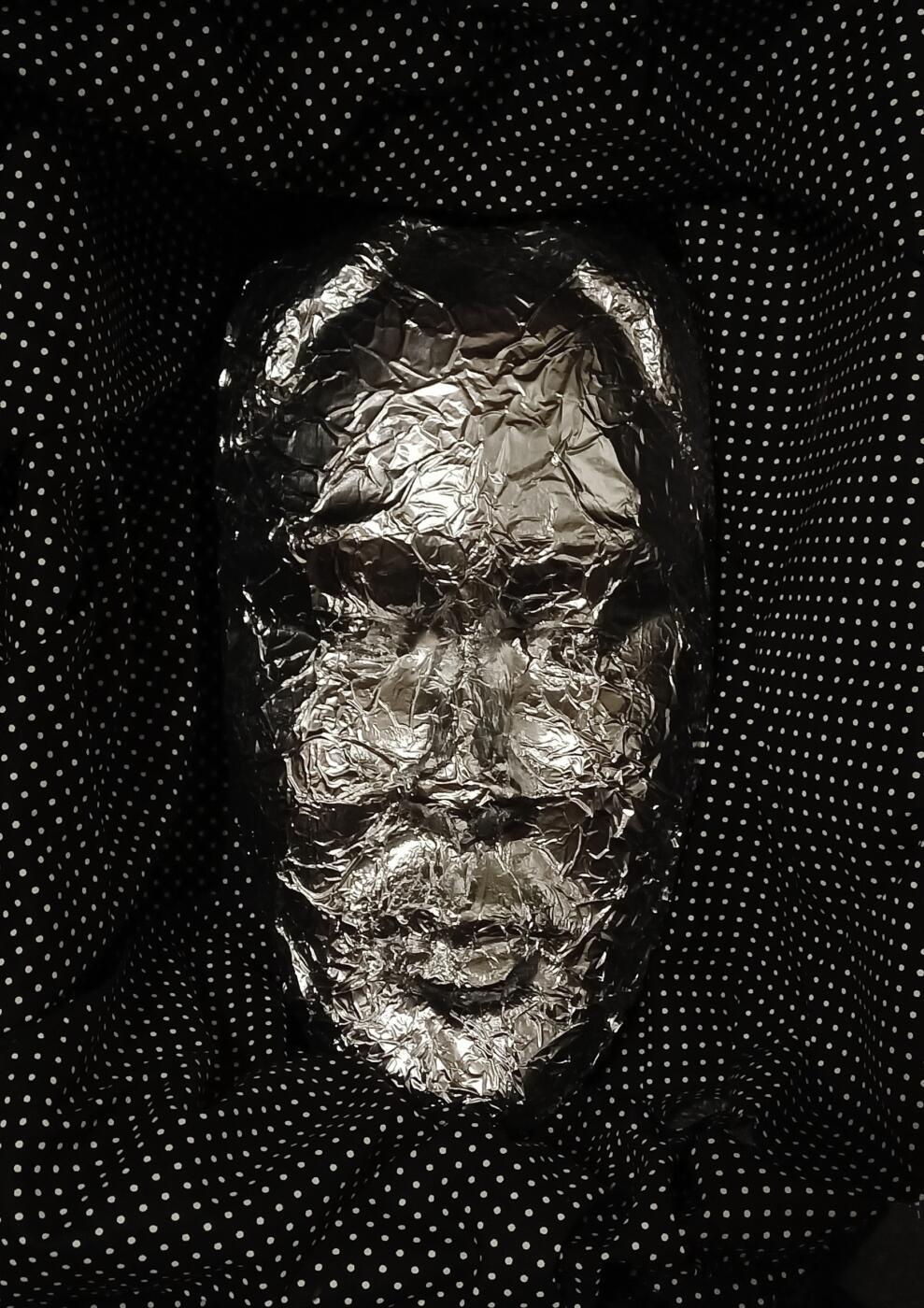 In the center of the image is a mask made of shiny, tin foil like material. Surrounding the mask is a black background with white polka dots.