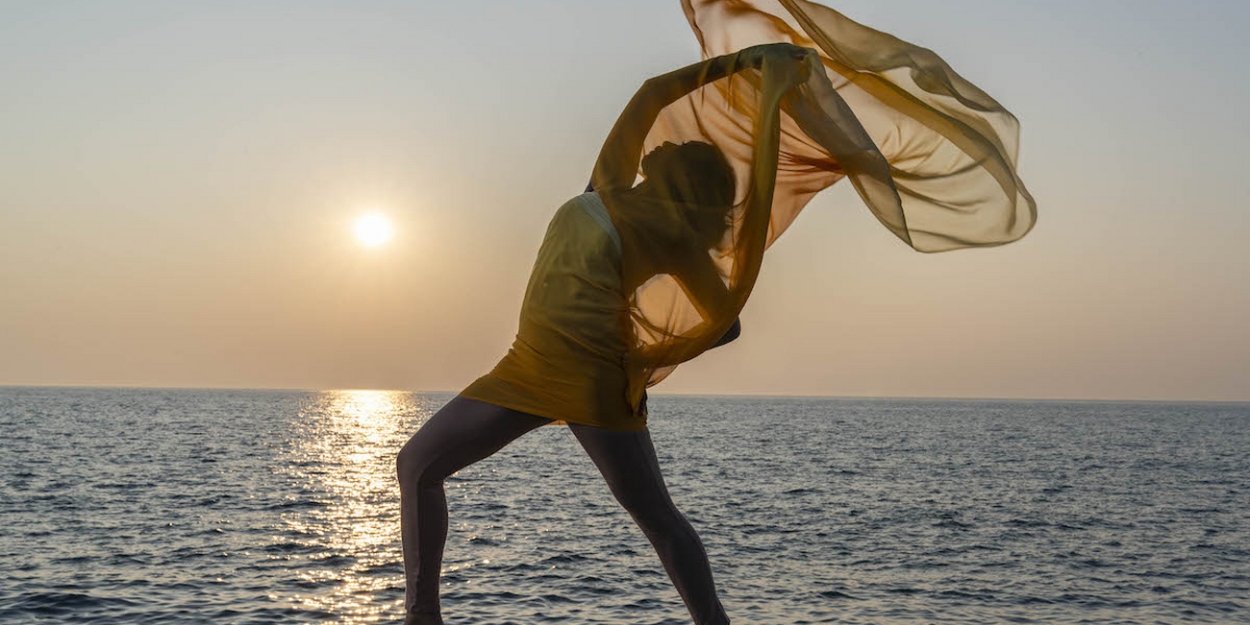 Image of a person mid-movement with a yellow garment splayed out in the wind. The lake fills the background and the rising sun creates an angelic-like light in the image.