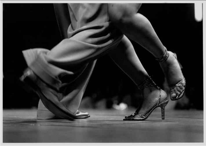 Image of two dancers' legs. They appear mid-movement wearing sparkly high heels and leather dress shoes.