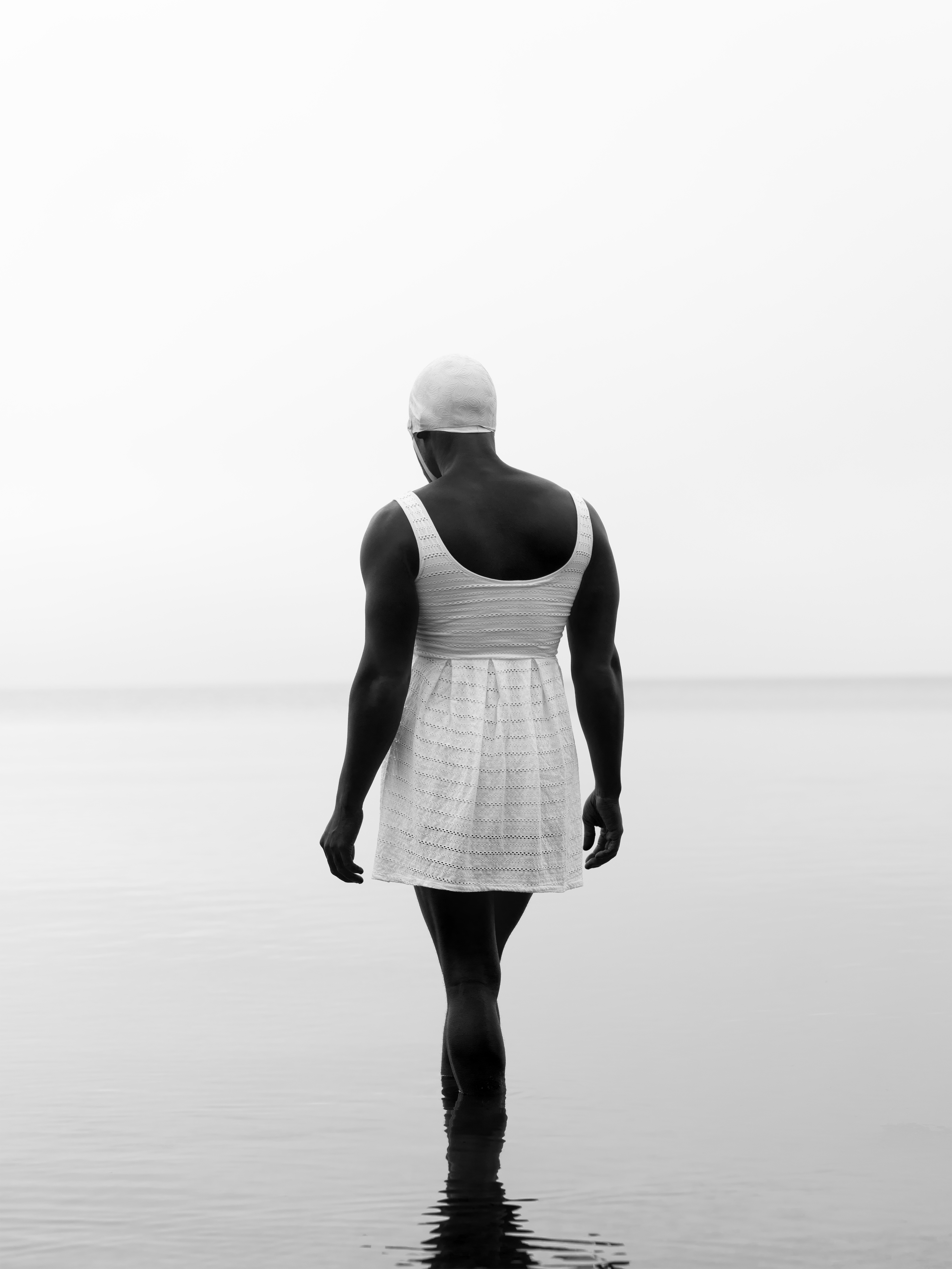  a black figure on shoaled beach stands with their back to the viewer. They are wearing a white swim dress and cap. Underneath them the water is still, and sky is clear. The ripple of their shadow trails off into the bottom of the image. 