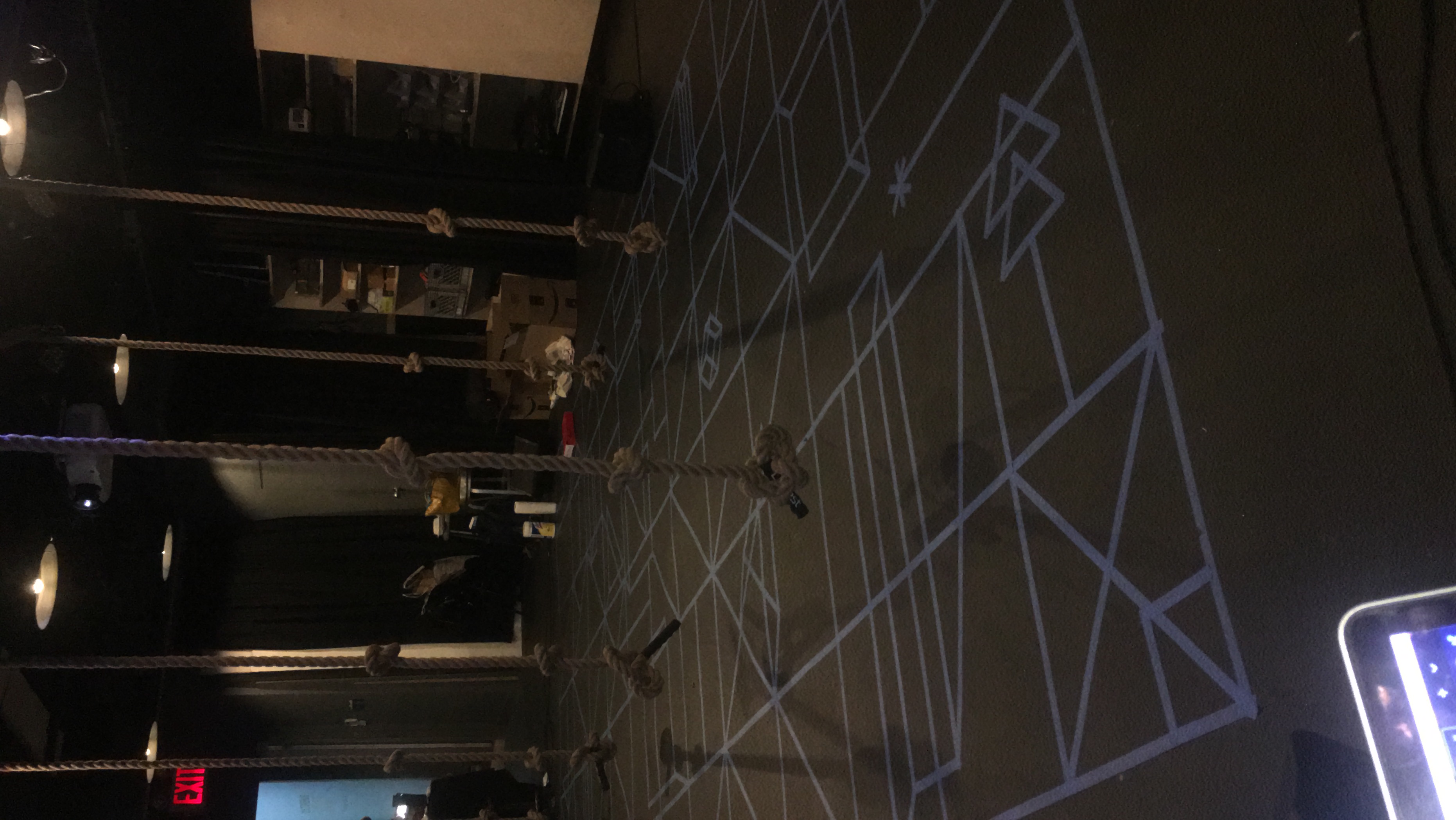  Photo. Small performance space, walls and floor painted black. In center is a blue floor design based in rhythmmetrics, sacred geometry, and hop scotch. Hanging from the cieling, over the floor design are 5 knotted climbing ropes. 