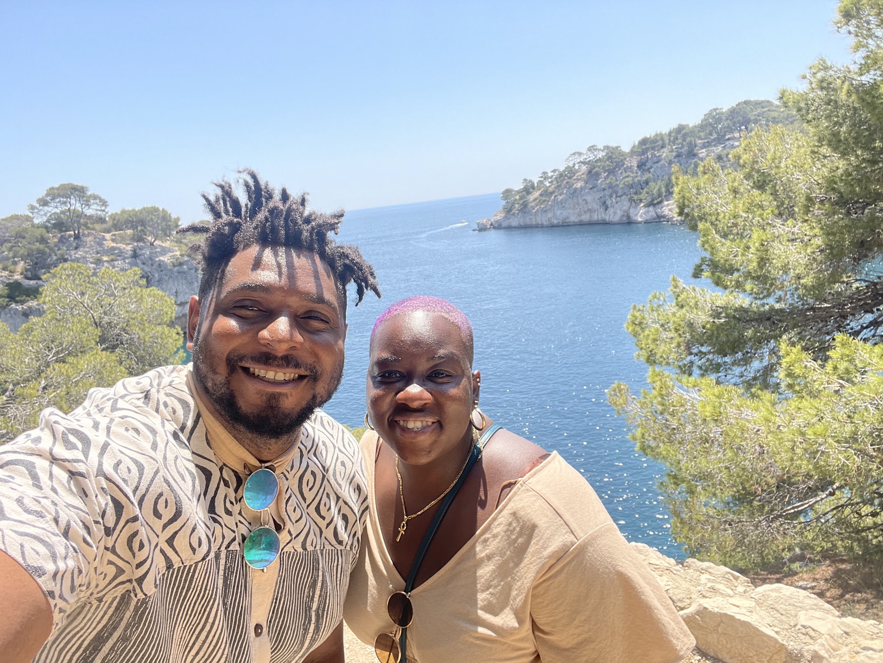  Two smiley, Black folks, taking a selfie. Cliff recede in the background, as foliage wraps the edges of the photograph. Directly behind them sits a very blue body of water, the Mediterranean Ocean. 