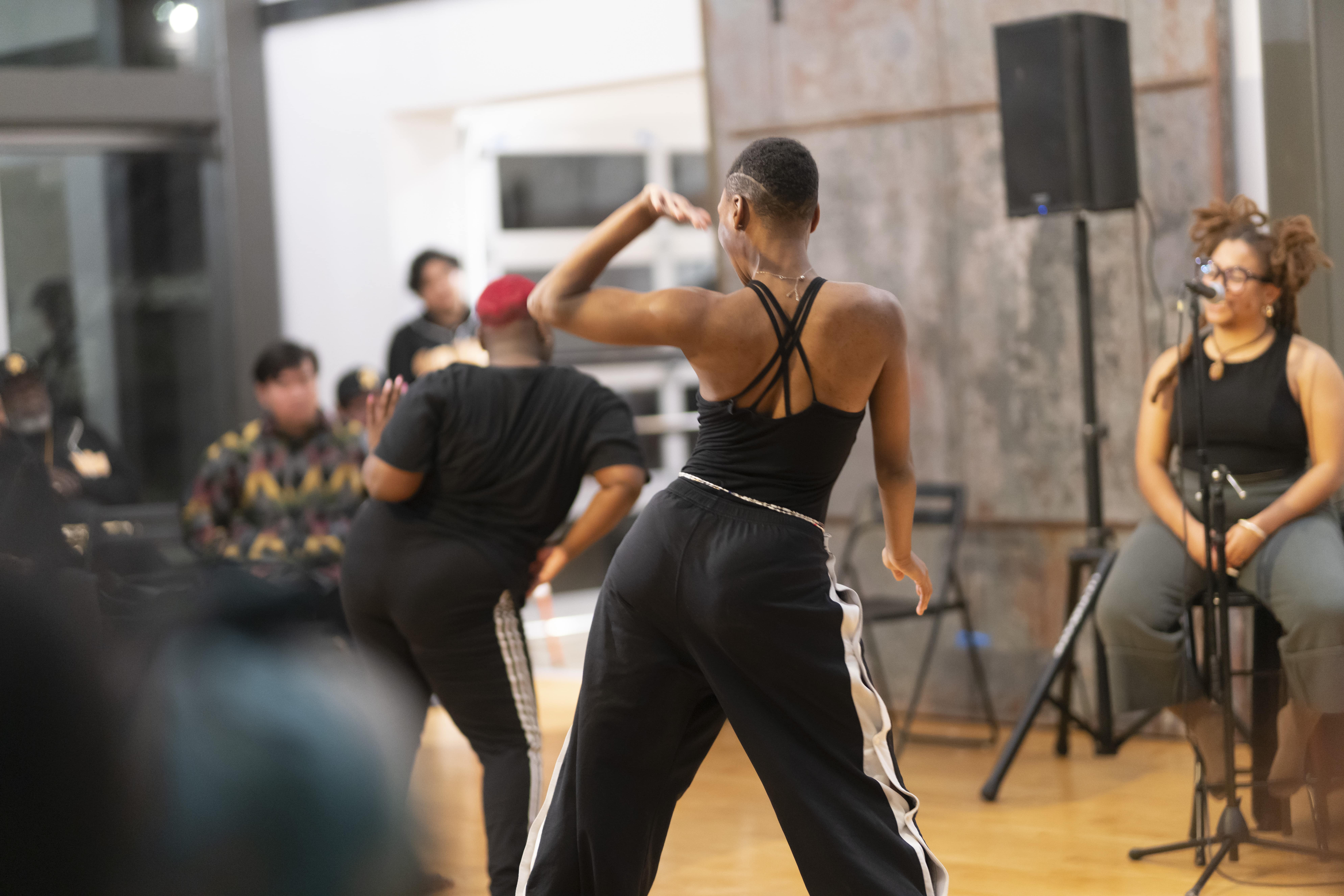  Deja Hood (at the foreground) & Keyierra Collins (in the background) perform at rehearse, play, jubilate at Dorchester Art + Housing Collaborative. Aaliyah Christina sits on a stool and speaks into a mic (blurred in the background). 