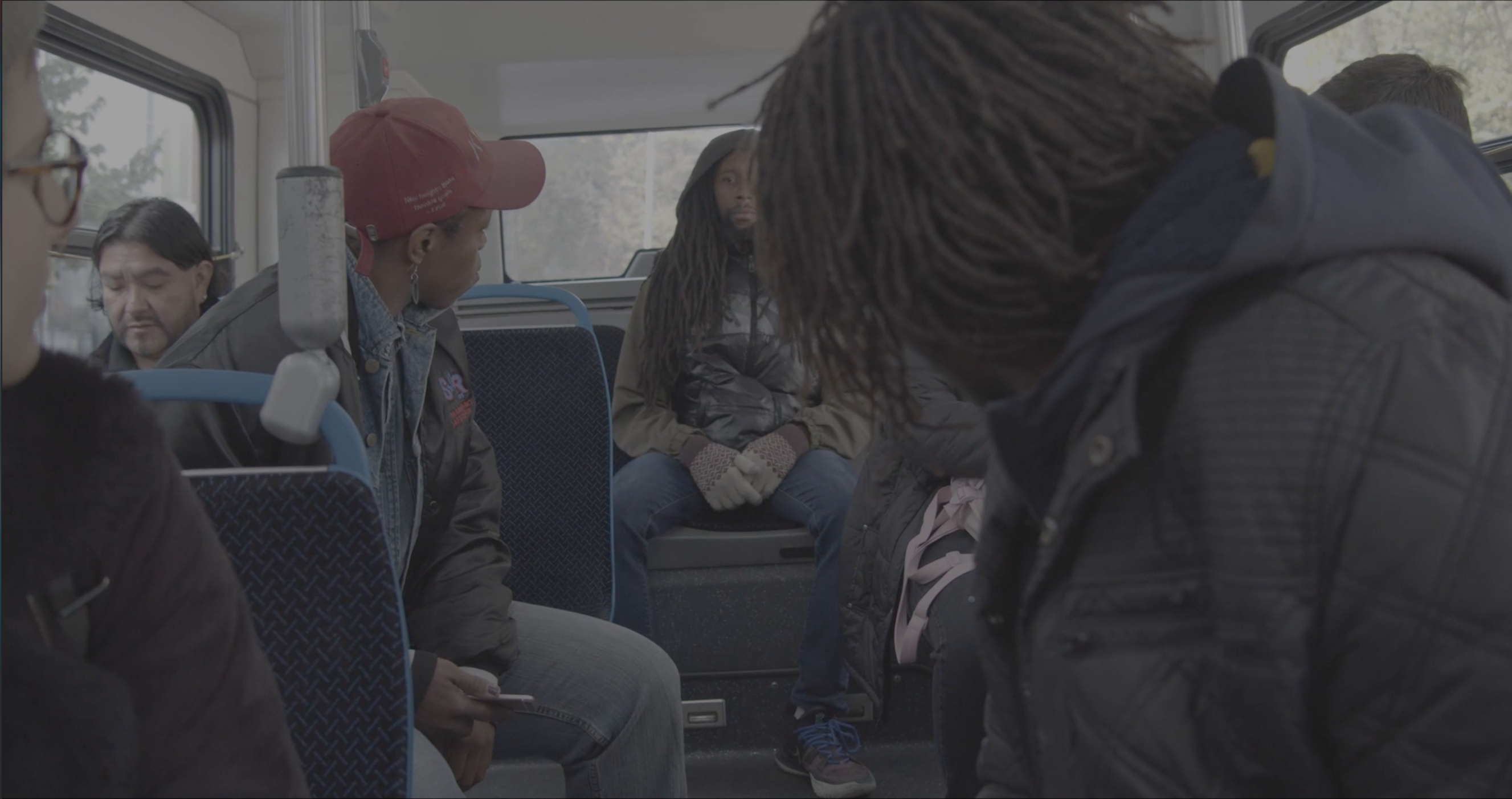  An ethnically diverse group of passengers are sitting on a bus, with their body partially turned around and looking at a young Black man, sitting down in the middle of the last row of seats. Everyone in the scene is wearing denim and "everyday" clothes. 