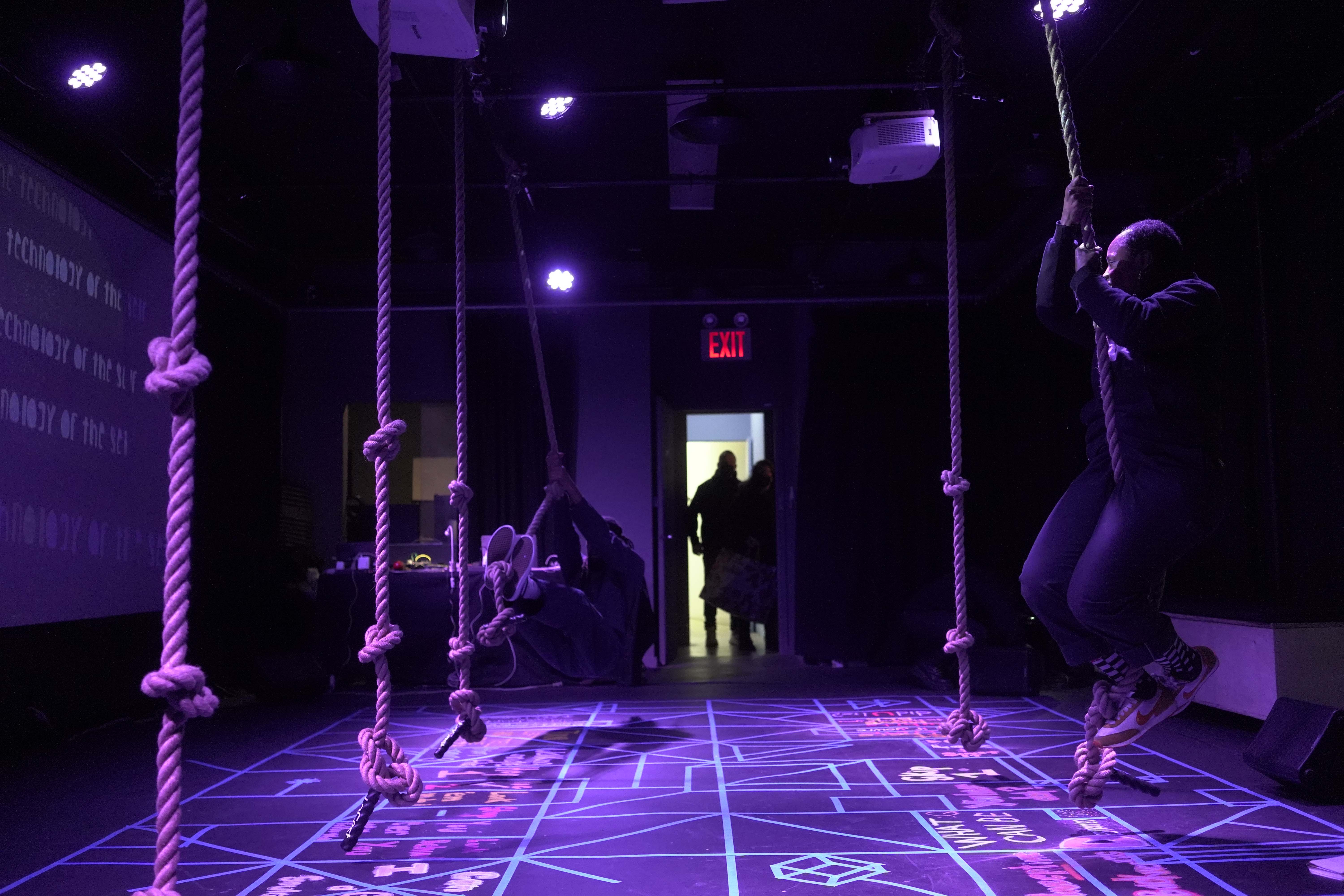  two black people, wearing dark blue coveralls, are swinging from ropes, over an interactive floor design,  done with a variety of bright colored chalk, and blue painters tape 