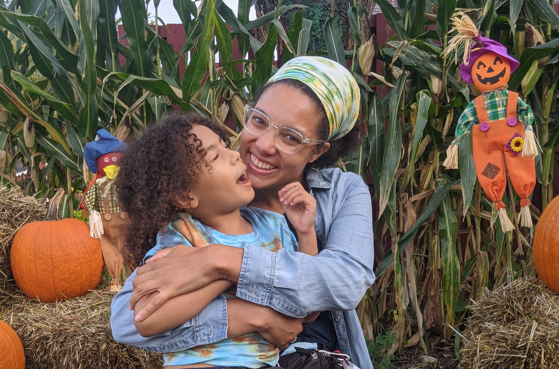  Light-skinned black mother wearing hand-dyed bandana and clear framed glasses with a big smile, holding her 6 yr old light-skinned kid with long brown curly hair in front of an array of fall items, corn stalks, hay bales, and pumpkins 